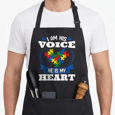 His autism acceptance apron - Puzzle heart in blue red yellow green with text I AM HIS VOICE HE IS MY HEART — shown on a black cotton canvas apron against a white background