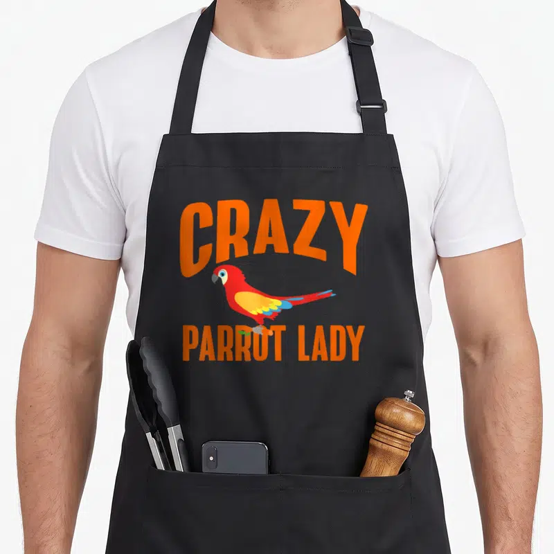 Wild parrot lady apron - Cartoon red parrot with orange CRAZY PARROT LADY text and blue yellow wings — shown on a black cotton canvas apron against a white background