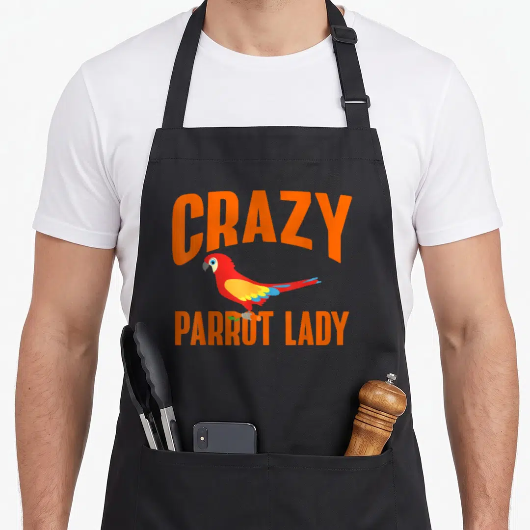 Wild parrot lady apron - Cartoon red parrot with orange CRAZY PARROT LADY text and blue yellow wings — shown on a black cotton canvas apron against a white background