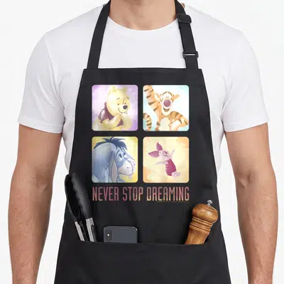 Stop novelty apron - Four watercolor portraits of Winnie the Pooh characters in a grid with NEVER STOP DREAMING text — shown on a black cotton canvas apron against a white background