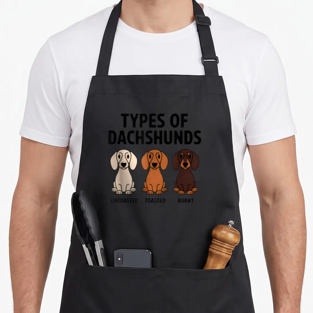 Wiener sausage apron - Three dachshunds in cream tan and brown labeled UNTOASTED TOASTED BURNT under TYPES — shown on a black cotton canvas apron against a white background