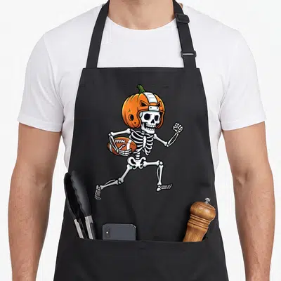 Spooky skeleton apron - Cartoon skeleton running with football wearing orange pumpkin helmet — shown on a black cotton canvas apron against a white background