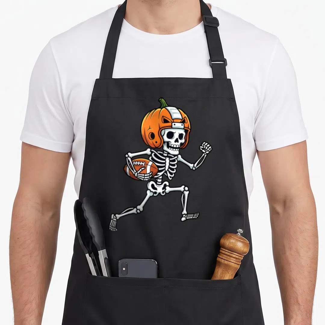 Spooky skeleton apron - Cartoon skeleton running with football wearing orange pumpkin helmet — shown on a black cotton canvas apron against a white background