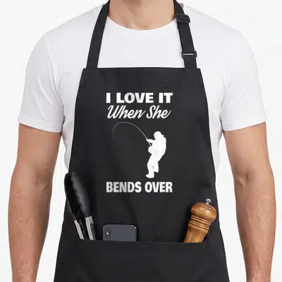 Vintage she fishing aprons - White text I Love It When She Bends Over with angler silhouette — shown on a black cotton canvas apron against a white background