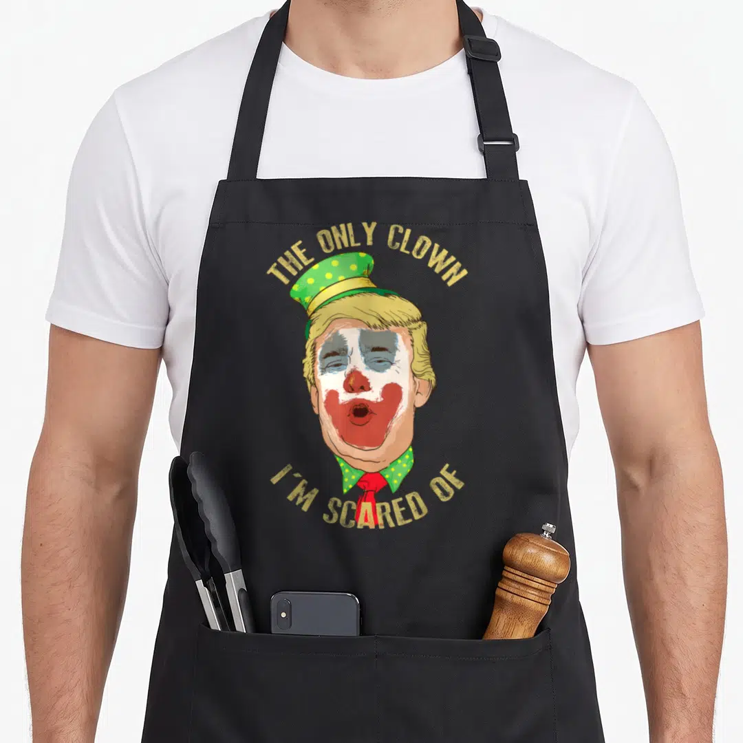 Scared clown aprons - Cartoon caricature of Donald Trump with polka-dot hat and red nose surrounded by gold text — shown on a black cotton canvas apron against a white background