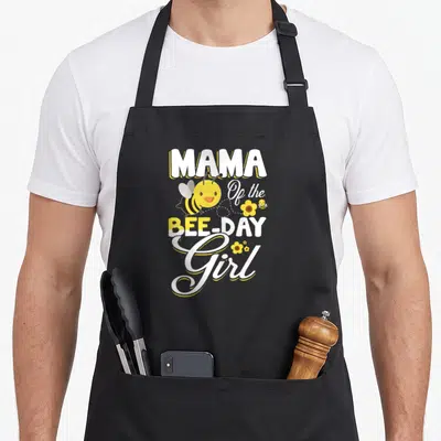 Mama bee aprons - Stacked Mama Bee Day Girl text with cartoon bees and yellow flowers — shown on a black cotton canvas apron against a white background