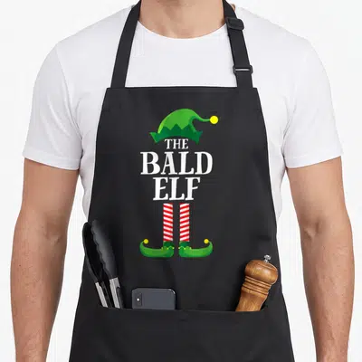 Bald christmas elf apron - THE BALD ELF text with green hat and red white striped legs — shown on a black cotton canvas apron against a white background