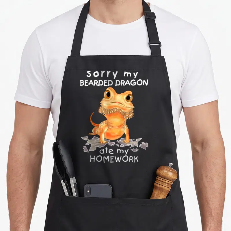 Lizard dragon apron - Orange cartoon bearded dragon standing on torn paper with white text — shown on a black cotton canvas apron against a white background