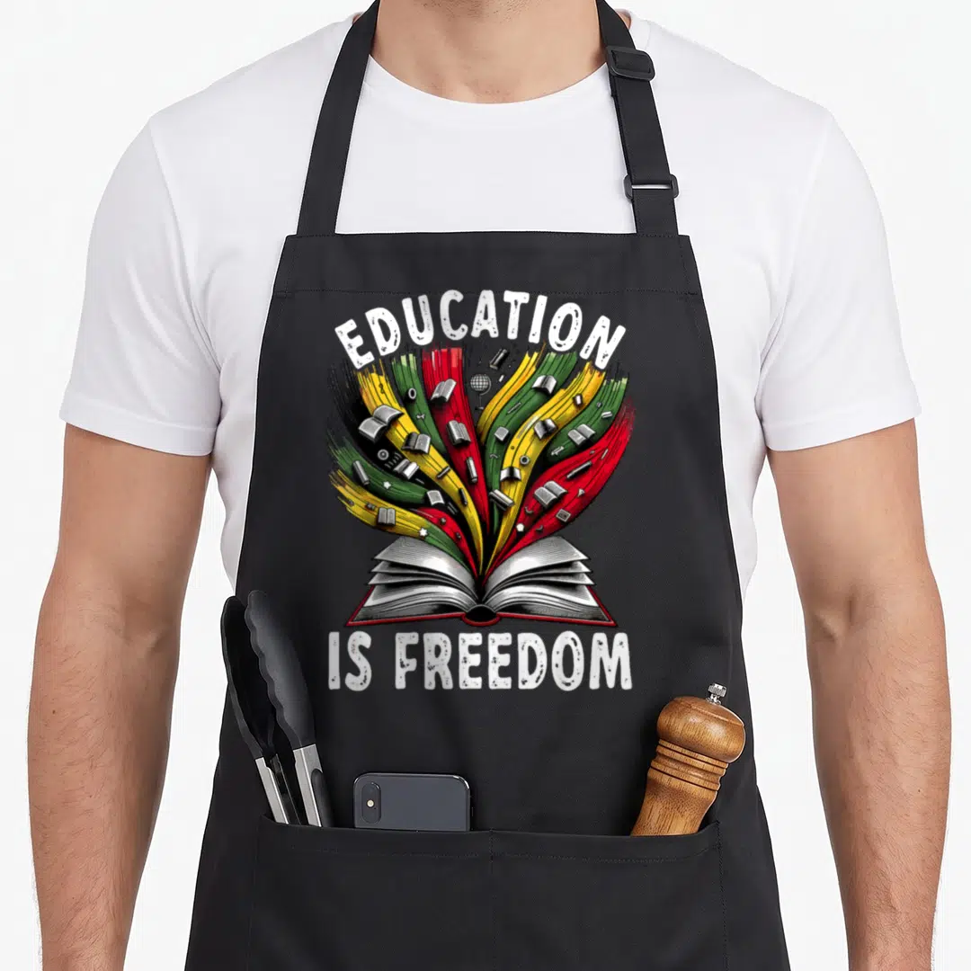 History education apron - Open book with red yellow green black streams and school supplies under EDUCATION IS FREEDOM — shown on a black cotton canvas apron against a white background