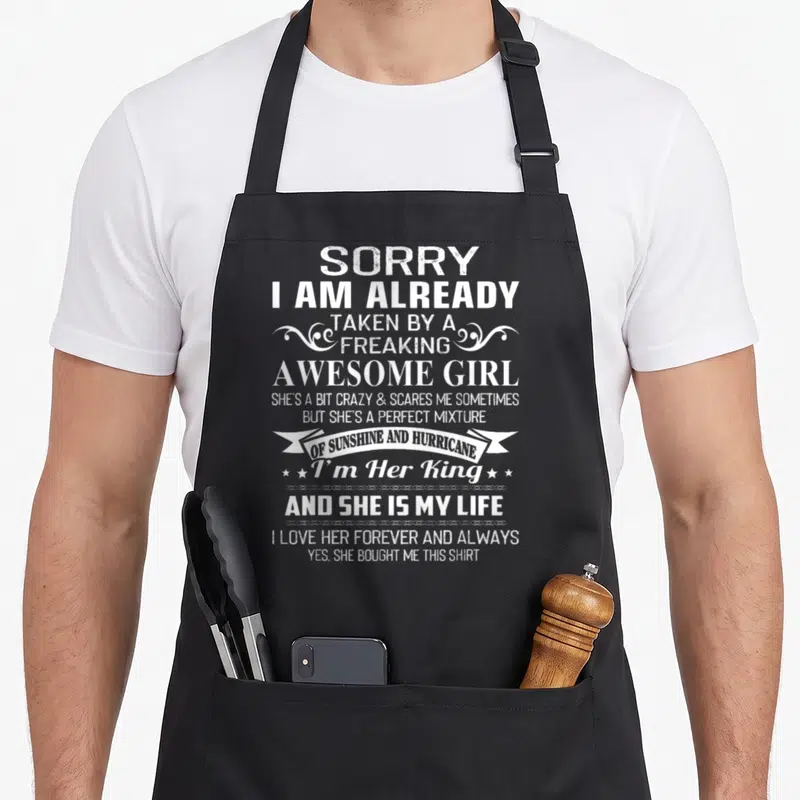 Already freaking apron - White distressed text quote about being taken by an awesome girl with banner and stars — shown on a black cotton canvas apron against a white background