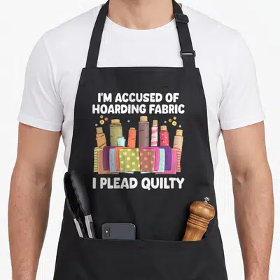 Sewer quilt apron - Cartoon fabric rolls and folded scraps with white text I'M ACCUSED OF HOARDING FABRIC I PLEAD — shown on a black cotton canvas apron against a white background
