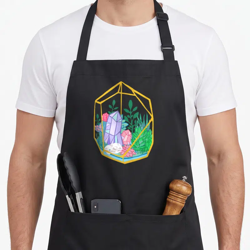 Terrarium goth apron - Cartoon terrarium with yellow frame, purple crystals, green succulents, and pink rocks — shown on a black cotton canvas apron against a white background