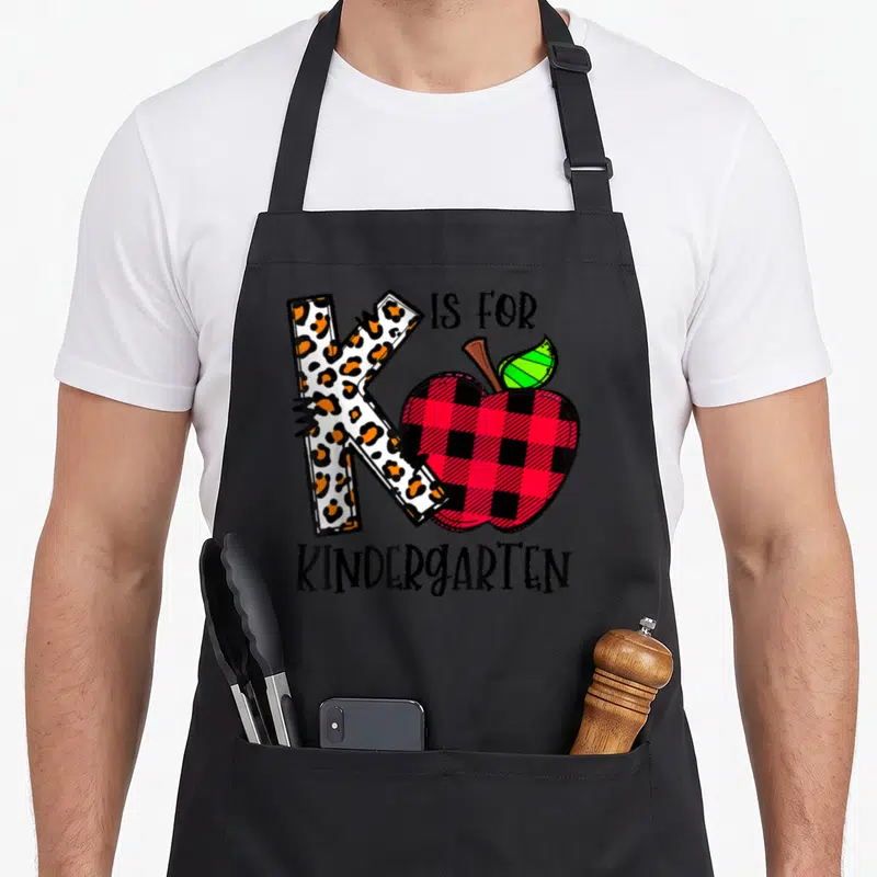 Funny leopard kindergarten teacher apron - Leopard print K and red buffalo plaid apple with IS FOR KINDERGARTEN text — shown on a black cotton canvas apron against a white background