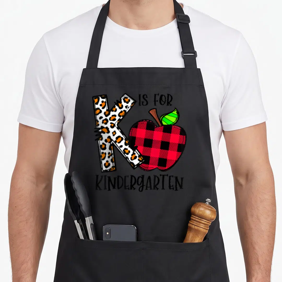 Funny leopard kindergarten teacher apron - Leopard print K and red buffalo plaid apple with IS FOR KINDERGARTEN text — shown on a black cotton canvas apron against a white background
