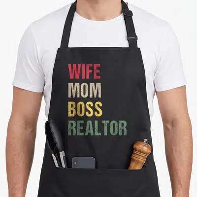 Boss wife apron - Stacked WIFE MOM BOSS REALTOR text in red beige yellow and green distressed fonts — shown on a black cotton canvas apron against a white background
