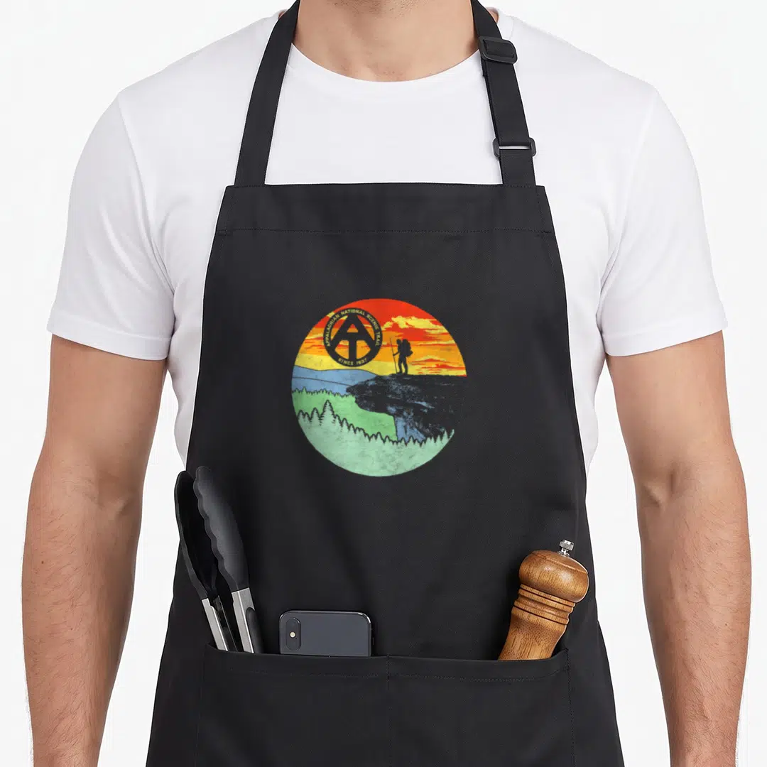 Appalachian camp trailer life apron - Retro circular hiker on cliff with Appalachian National Scenic Trail text — shown on a black cotton canvas apron against a white background