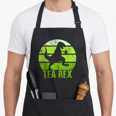 Rex tea party aprons - Grey T-Rex silhouette holding a cup inside a lime green striped circle with TEA REX text below — shown on a black cotton canvas apron against a white background