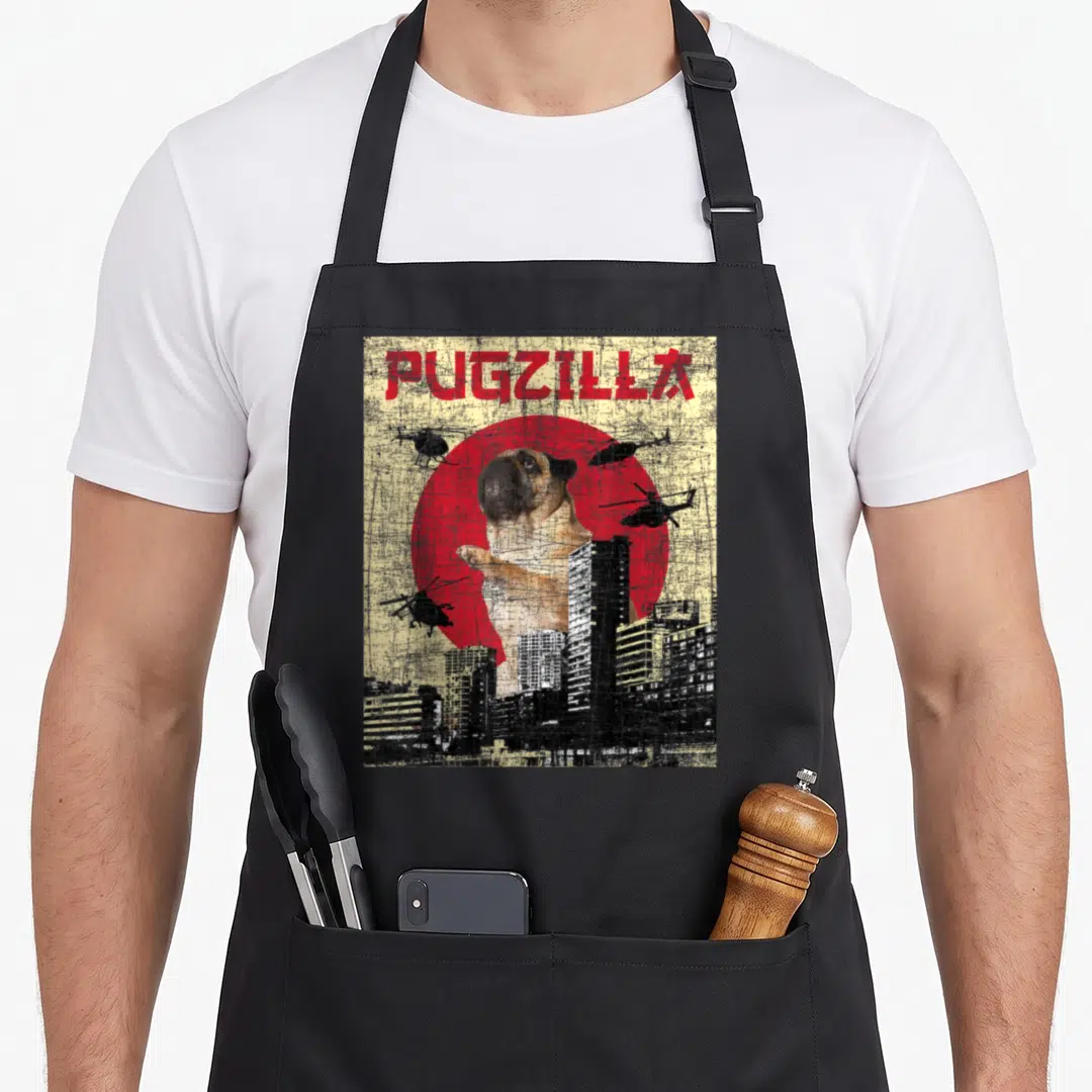 Pugzilla dog trainer apron - Giant pug behind city skyline with helicopters flying near red circle and distressed text — shown on a black cotton canvas apron against a white background