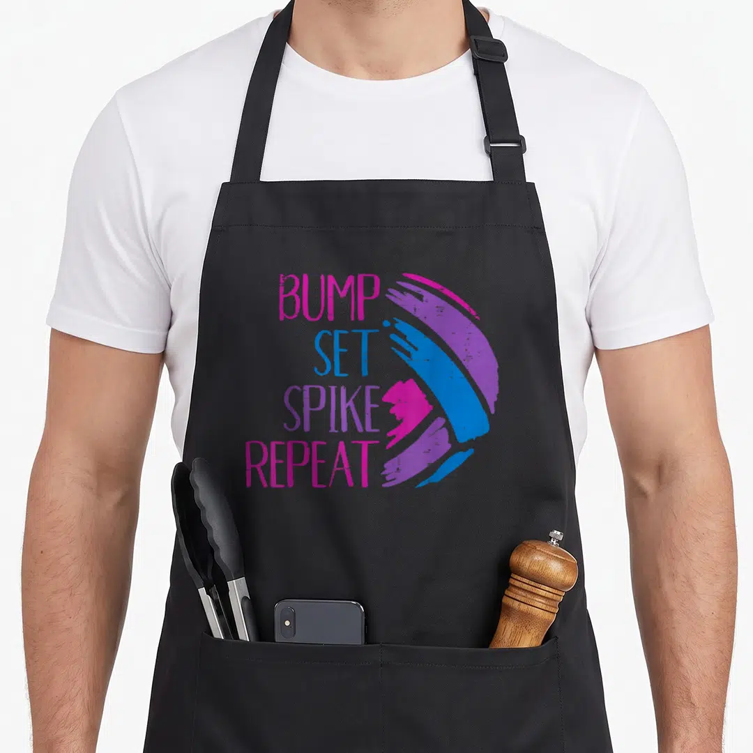 Bump spiker apron - Stacked BUMP SET SPIKE REPEAT text next to a multi-colored brushstroke volleyball illustration — shown on a black cotton canvas apron against a white background