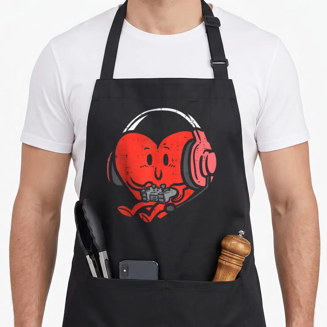 Youth valentine's day apron - Red cartoon heart wearing gaming headset and holding grey controller with small legs — shown on a black cotton canvas apron against a white background