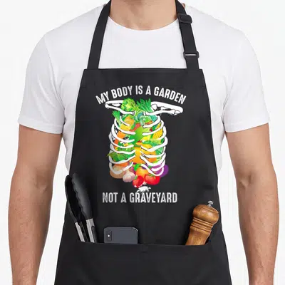 Body veggie apron - Cartoon ribcage filled with vegetables and text My Body Is A Garden Not A Graveyard — shown on a black cotton canvas apron against a white background