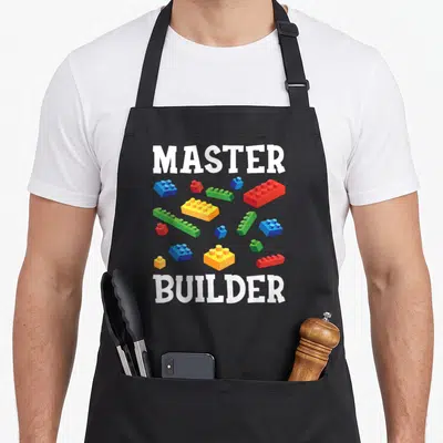 Block building grill master apron - White MASTER and BUILDER text with scattered red blue yellow green toy bricks — shown on a black cotton canvas apron against a white background