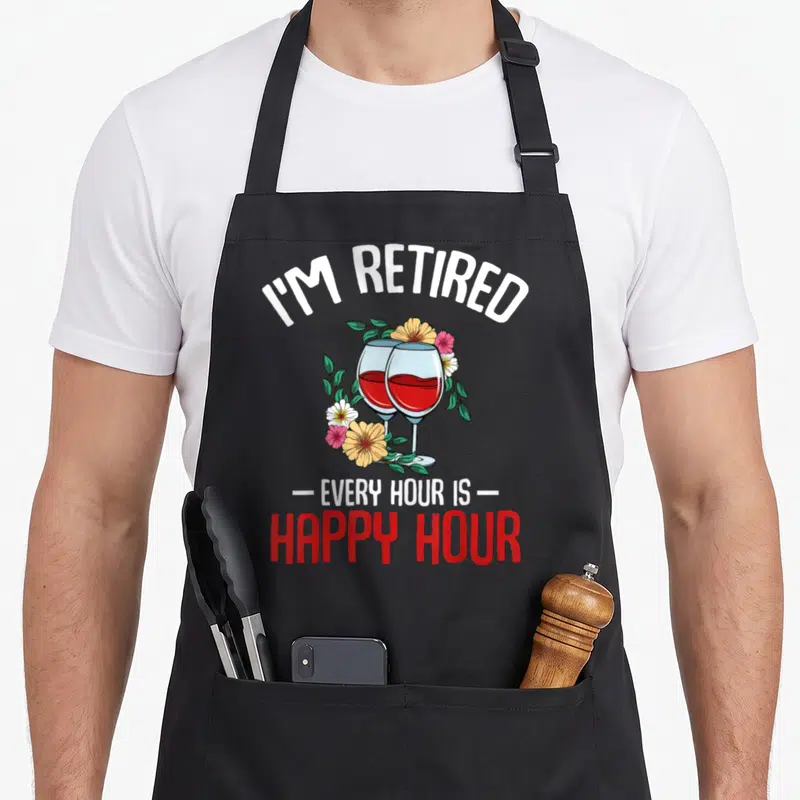 Hour retiree apron - Two wine glasses with red liquid surrounded by yellow, green, white, and pink flowers — shown on a black cotton canvas apron against a white background