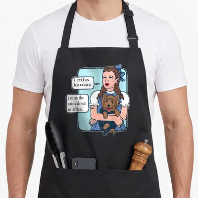 Miss joke apron - Cartoon girl holding brown dog, blue frame, text i miss kansas and rains down in africa — shown on a black cotton canvas apron against a white background
