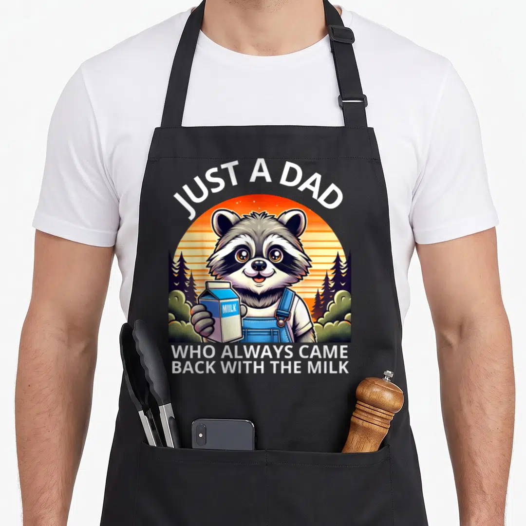 Cute raccoon milk aprons - Cartoon raccoon in blue overalls holding a milk carton under arched text Just a Dad — shown on a black cotton canvas apron against a white background