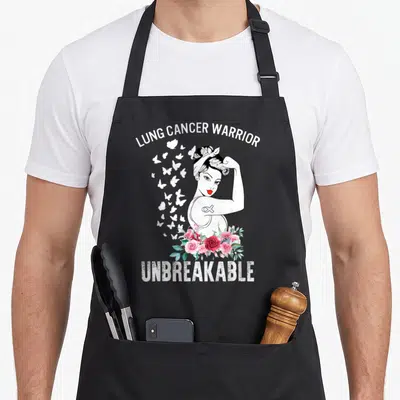 Unbreakable lungs apron - Cartoon woman flexing arm with LUNG CANCER WARRIOR text, butterflies, and pink roses — shown on a black cotton canvas apron against a white background