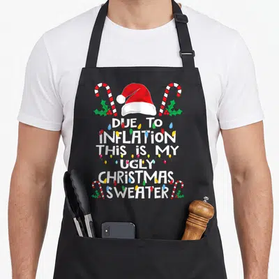 Due ugly christmas sweater apron - Stacked white text DUE TO INFLATION THIS IS MY UGLY CHRISTMAS SWEATER with Santa hat — shown on a black cotton canvas apron against a white background