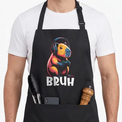 Bruh capybara video game apron - Cartoon capybara wearing blue headphones and holding a controller with white BRUH text — shown on a black cotton canvas apron against a white background