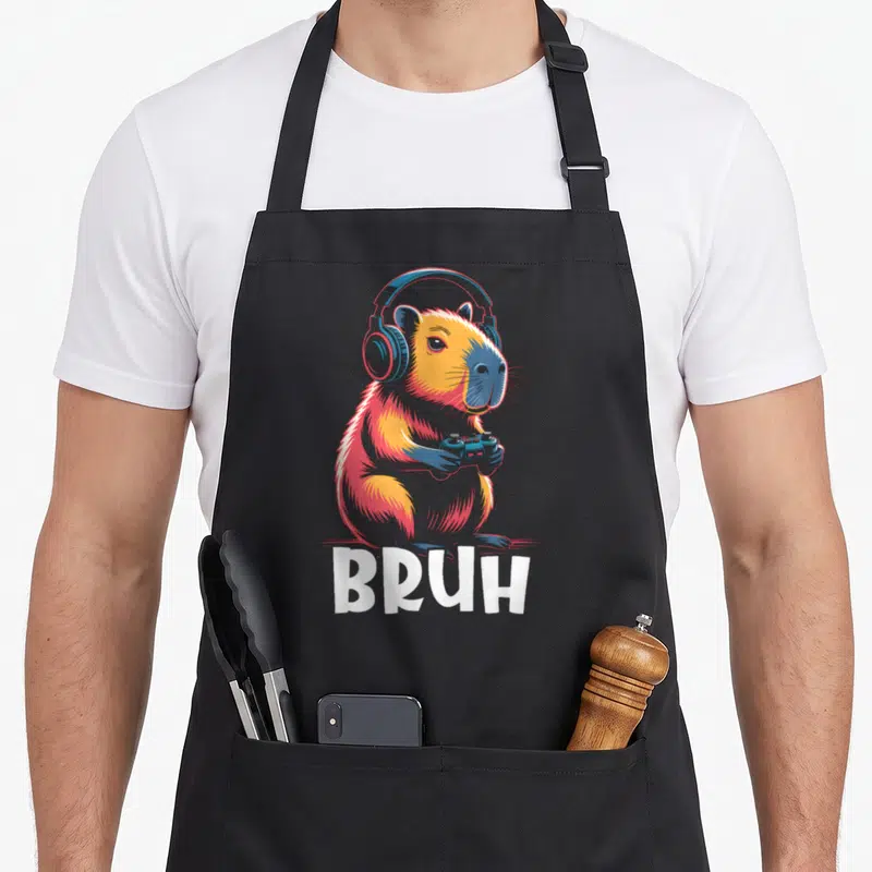 Bruh capybara video game apron - Cartoon capybara wearing blue headphones and holding a controller with white BRUH text — shown on a black cotton canvas apron against a white background