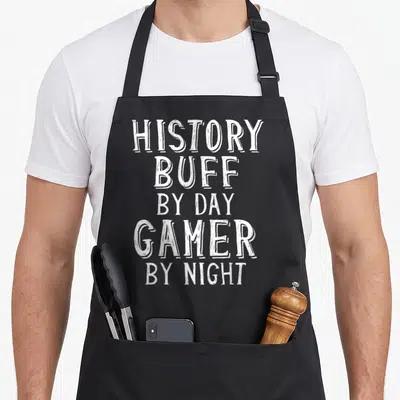 History gaming apron - White stacked typography reading History Buff By Day Gamer By Night on grey background — shown on a black cotton canvas apron against a white background