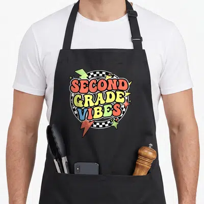 Groovy second 2nd apron - Stacked SECOND GRADE VIBES text over checkered circle with colorful lightning bolts and stars — shown on a black cotton canvas apron against a white background