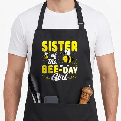 Sister hive bee aprons - Stacked yellow and white text reading Sister of the Bee Day Girl with two cartoon bees — shown on a black cotton canvas apron against a white background