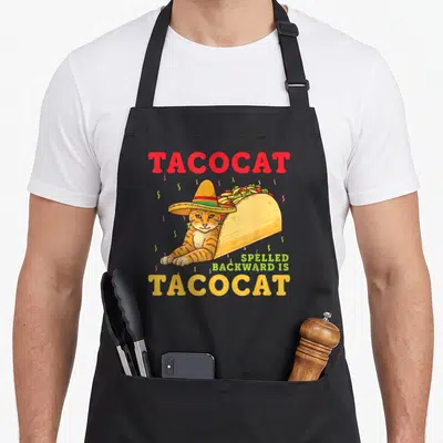 Spelled cat apron - TACOCAT text with sombrero cat in taco shell and confetti — shown on a black cotton canvas apron against a white background