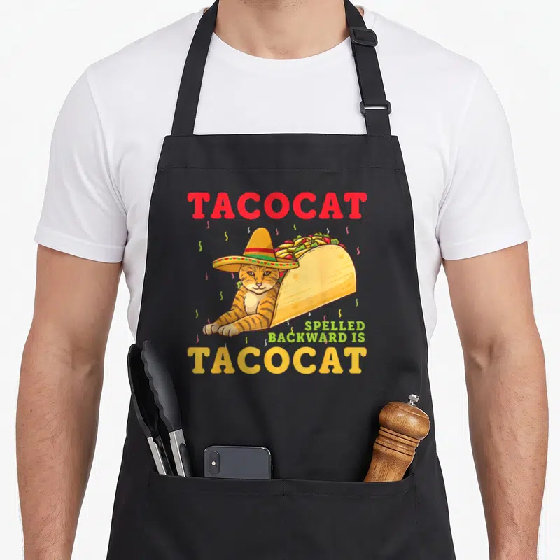 Spelled cat apron - TACOCAT text with sombrero cat in taco shell and confetti — shown on a black cotton canvas apron against a white background