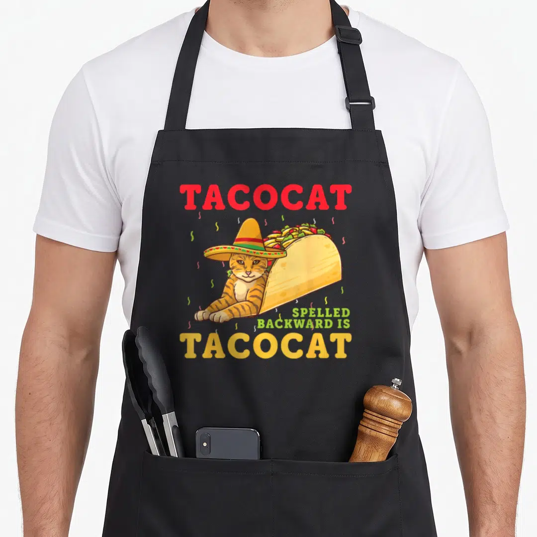 Spelled cat apron - TACOCAT text with sombrero cat in taco shell and confetti — shown on a black cotton canvas apron against a white background