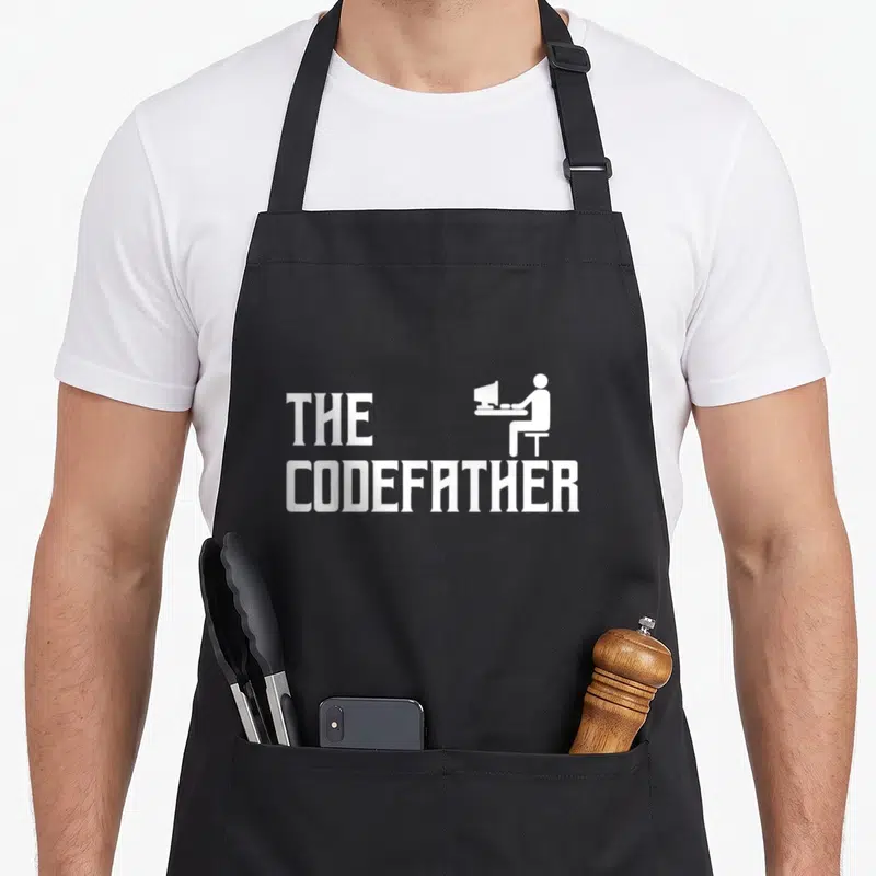 Codefather coder apron - White THE CODEFATHER text with sitting programmer silhouette at computer desk — shown on a black cotton canvas apron against a white background