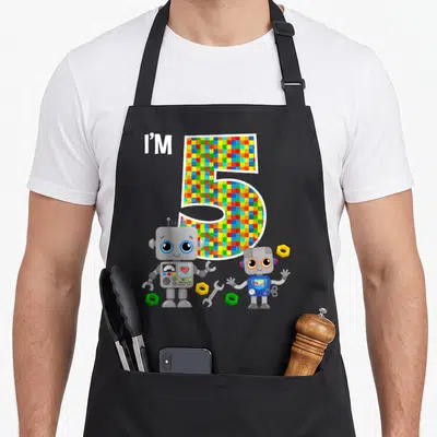 Robot 5th birthday apron - I'M 5 text with pixelated number, two cartoon robots, nuts and wrench on grey background — shown on a black cotton canvas apron against a white background