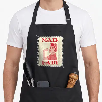 Stamp postal aprons - Red and beige stamp graphic showing a woman flexing arm with text mail lady — shown on a black cotton canvas apron against a white background