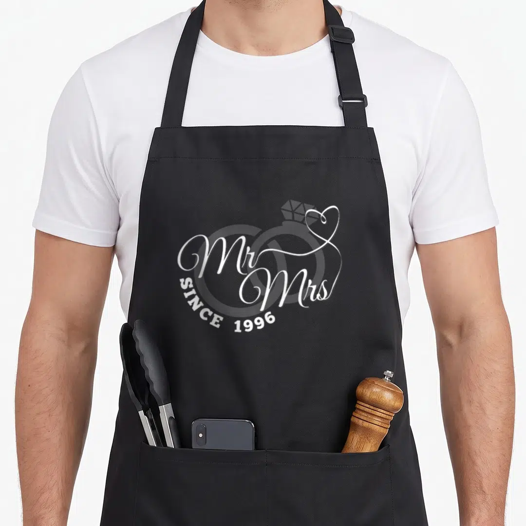 27th mr mrs aprons - White script reading Mr Mrs Since 1996 over two interlocking rings with a heart detail — shown on a black cotton canvas apron against a white background