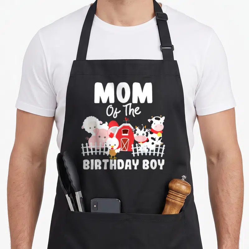 Birthday animal farm aprons - MOM Of The BIRTHDAY BOY text above cartoon sheep, pig, chicken, and cow by a red barn — shown on a black cotton canvas apron against a white background
