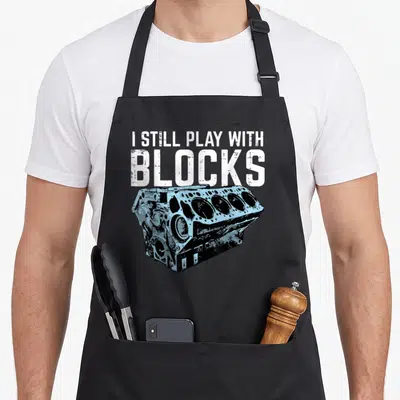 Mechanic car apron - I STILL PLAY WITH BLOCKS text above light blue engine block illustration with distressed white — shown on a black cotton canvas apron against a white background