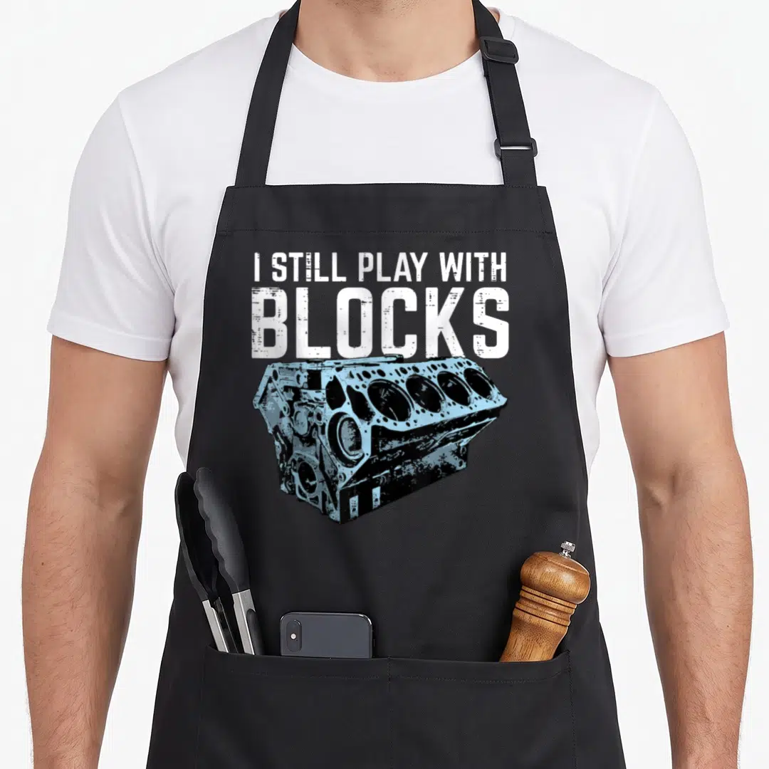Mechanic car apron - I STILL PLAY WITH BLOCKS text above light blue engine block illustration with distressed white — shown on a black cotton canvas apron against a white background