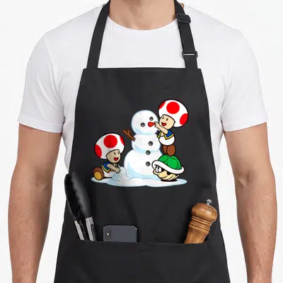 Snow toad apron - Cartoon snowman with two mushroom characters and green turtle shell using white red blue and green — shown on a black cotton canvas apron against a white background