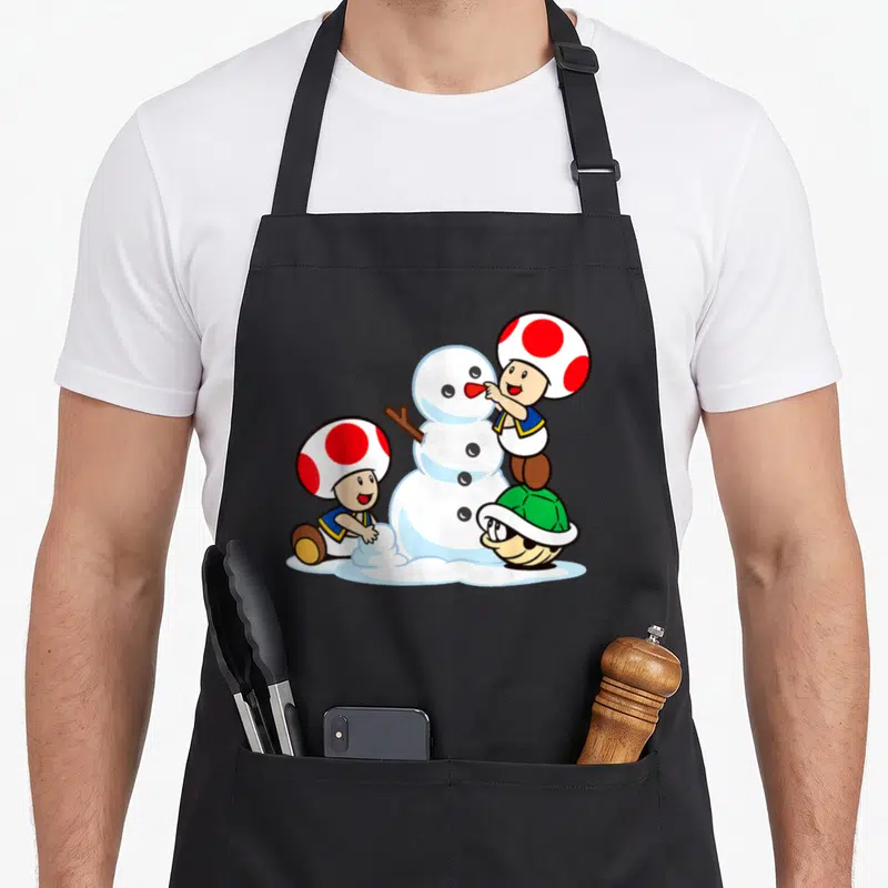 Snow toad apron - Cartoon snowman with two mushroom characters and green turtle shell using white red blue and green — shown on a black cotton canvas apron against a white background