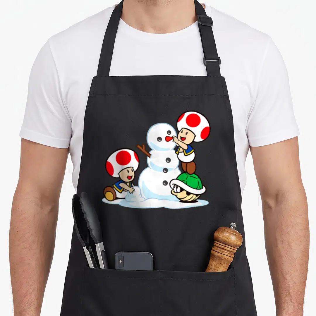 Snow toad apron - Cartoon snowman with two mushroom characters and green turtle shell using white red blue and green — shown on a black cotton canvas apron against a white background