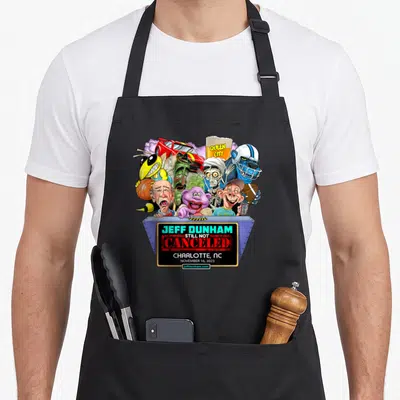 Dunham charlotte apron - Cartoon puppets above grey podium with Jeff Dunham Still Not Canceled text — shown on a black cotton canvas apron against a white background